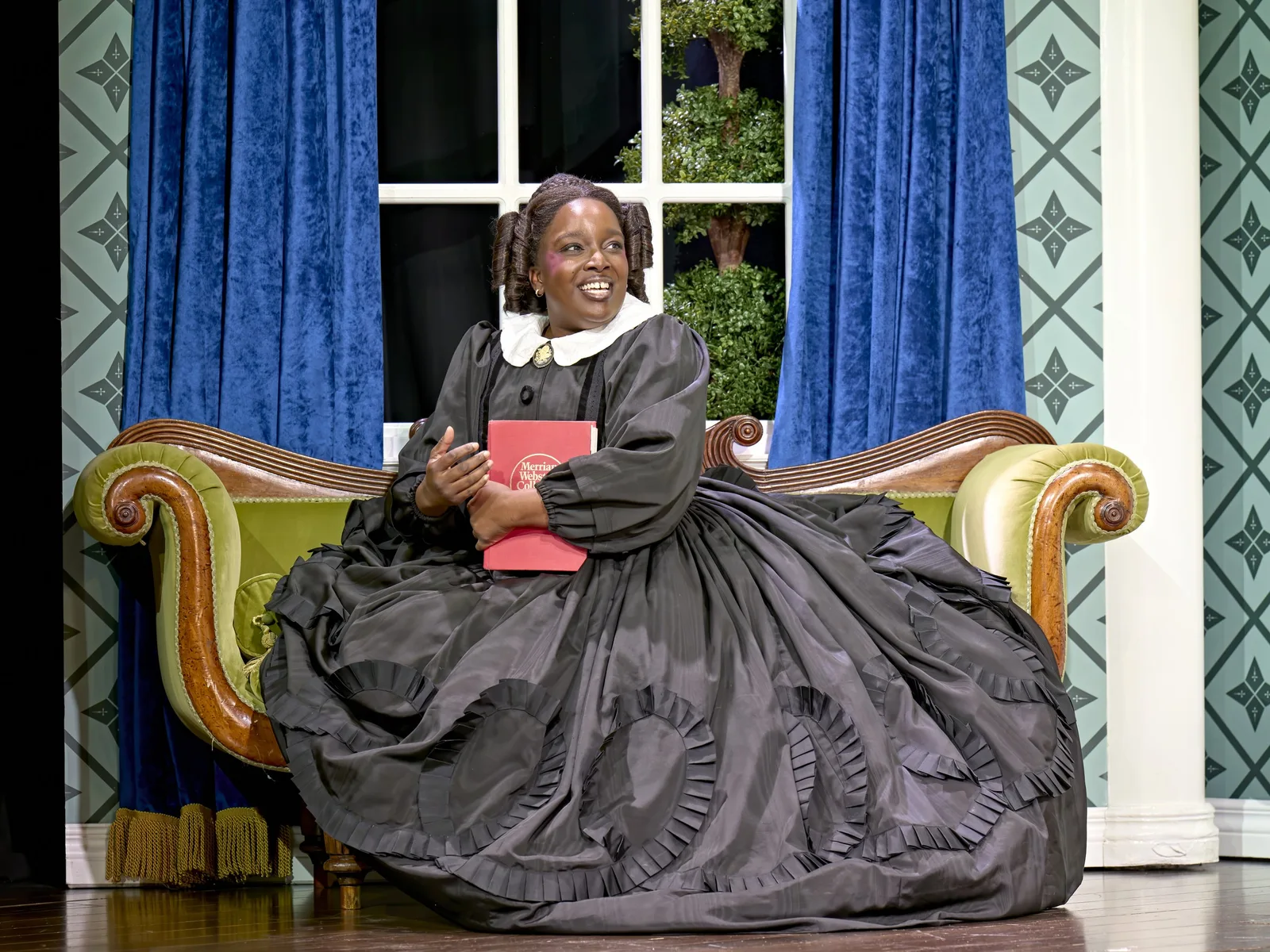 Lolly Adefope in a voluminous black Victorian gown and white collar sits on a curved green sofa beneath blue curtains, hugging a red book