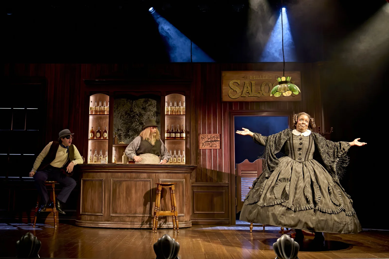 Lolly Adefope in a full grey crinoline gown flings her arms wide on a dimly lit stage beside a writing table and a framed portrait