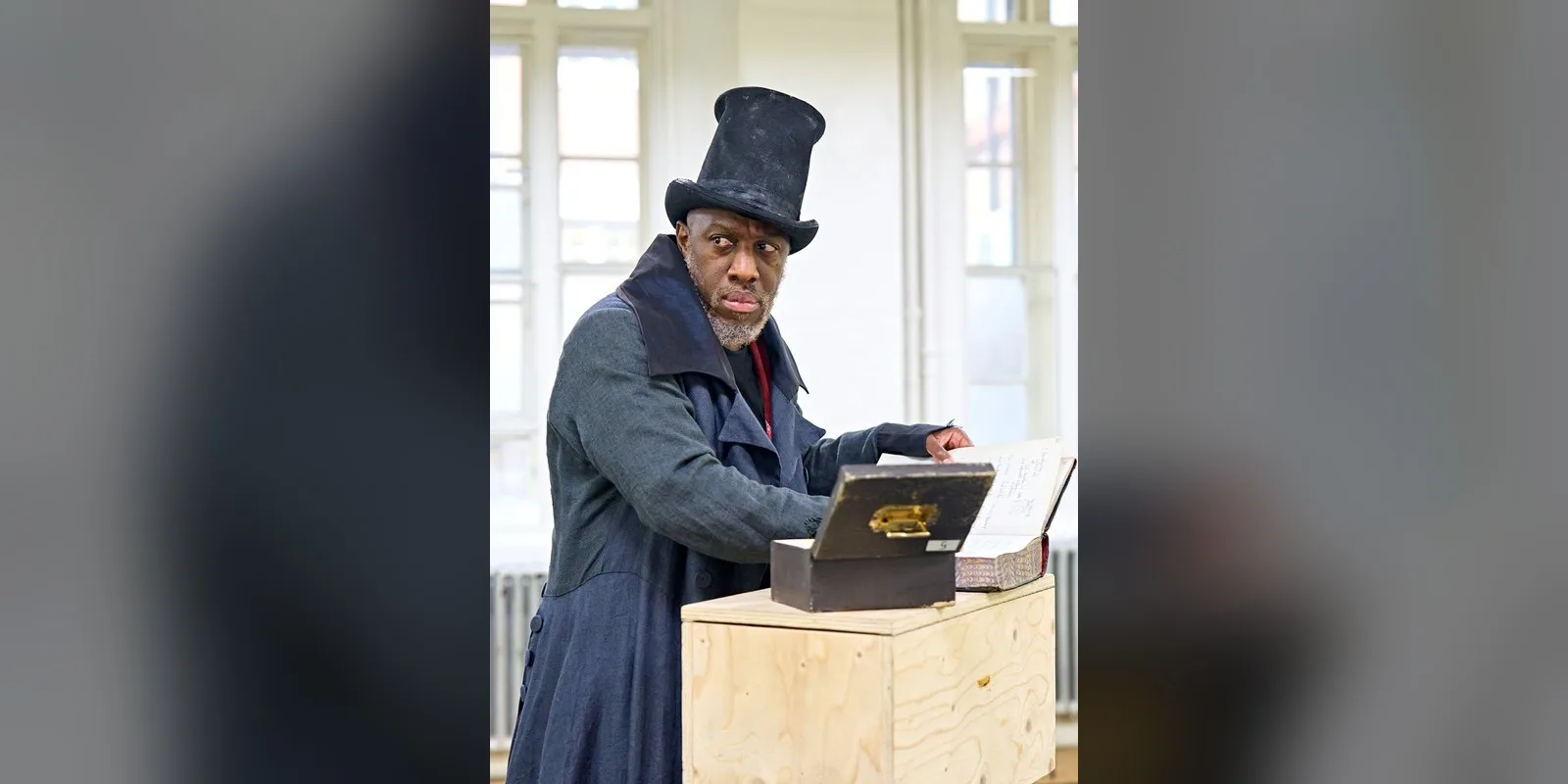An actor in a tall dark stovepipe hat and long dark coat leans over a small money box on a plain wooden crate in a bright rehearsal room