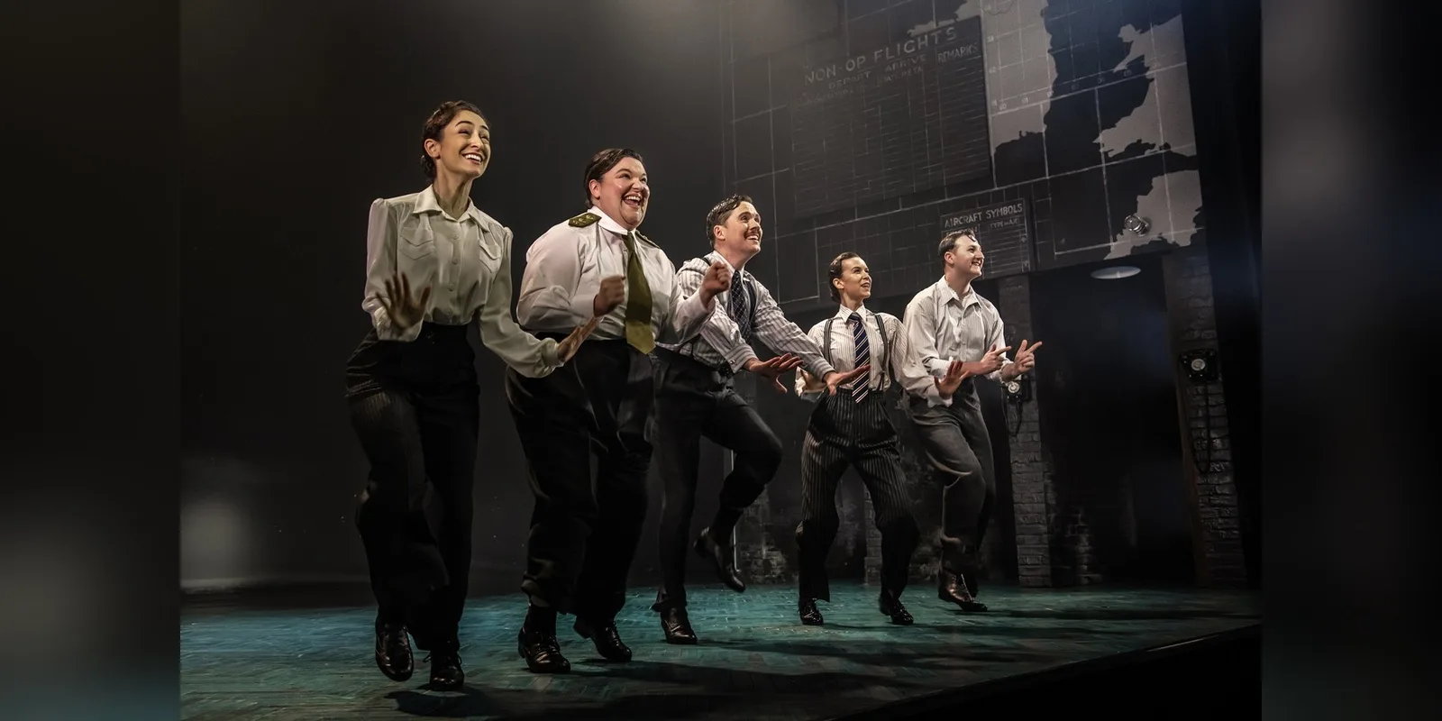 Five performers in 1940s shirts, ties and trousers dance in a line across the stage in front of a wartime map of Britain