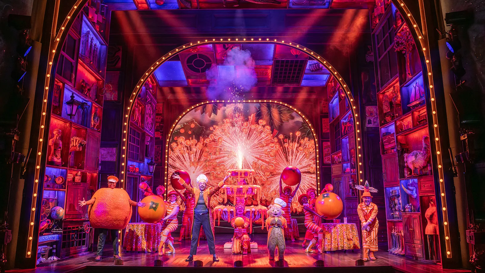 A brightly lit stage framed by a golden arch, with performers in colourful costumes and a small bear in a tall white cook's hat at the centre beneath a burst of sparkling fireworks