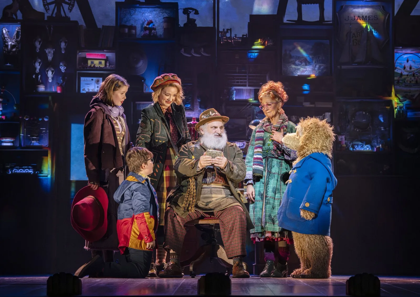 The bear in a blue duffel coat and red hat stands with a family and a white-bearded shopkeeper in a cluttered antiques shop