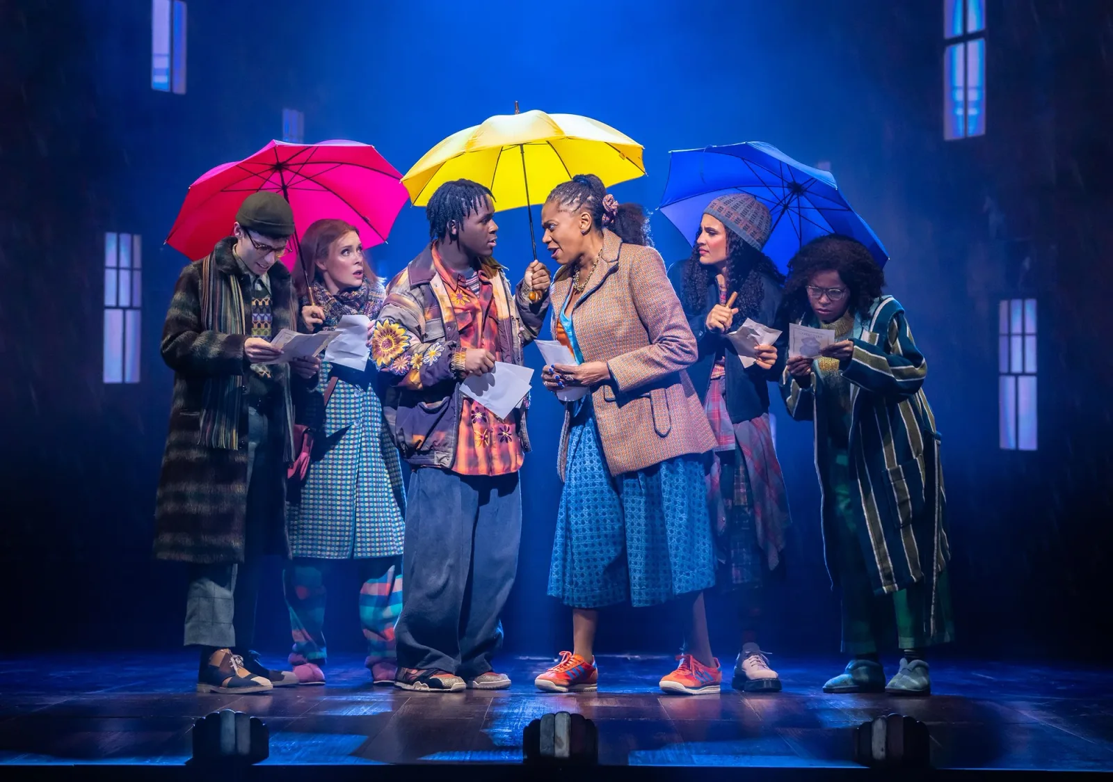 Performers in raincoats shelter under bright red, yellow and blue umbrellas on a rainy London street