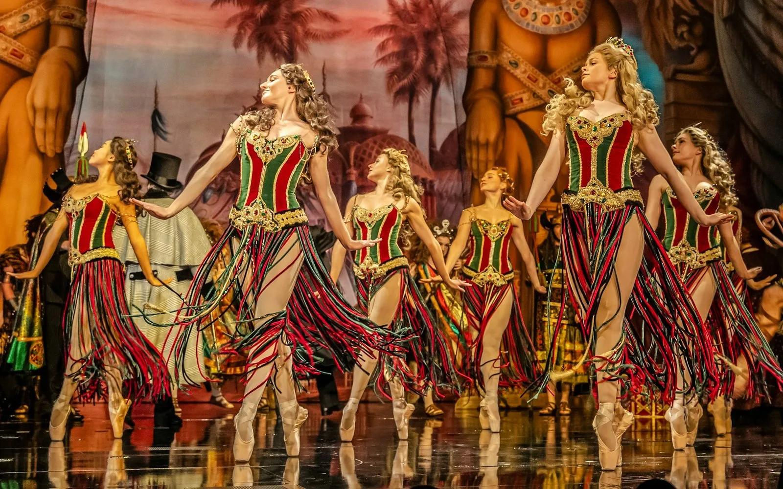 Ballet dancers in red and green striped costumes with long fringed skirts dancing in front of a painted backdrop in a scene from The Phantom of the Opera
