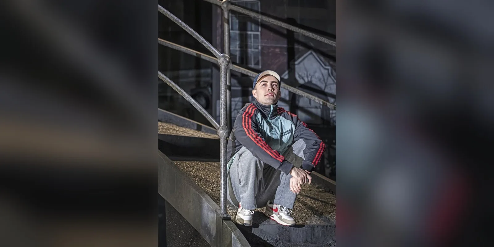 Elan Butler in a tracksuit and cap crouches on a metal staircase in the 2026 production of James Graham's play