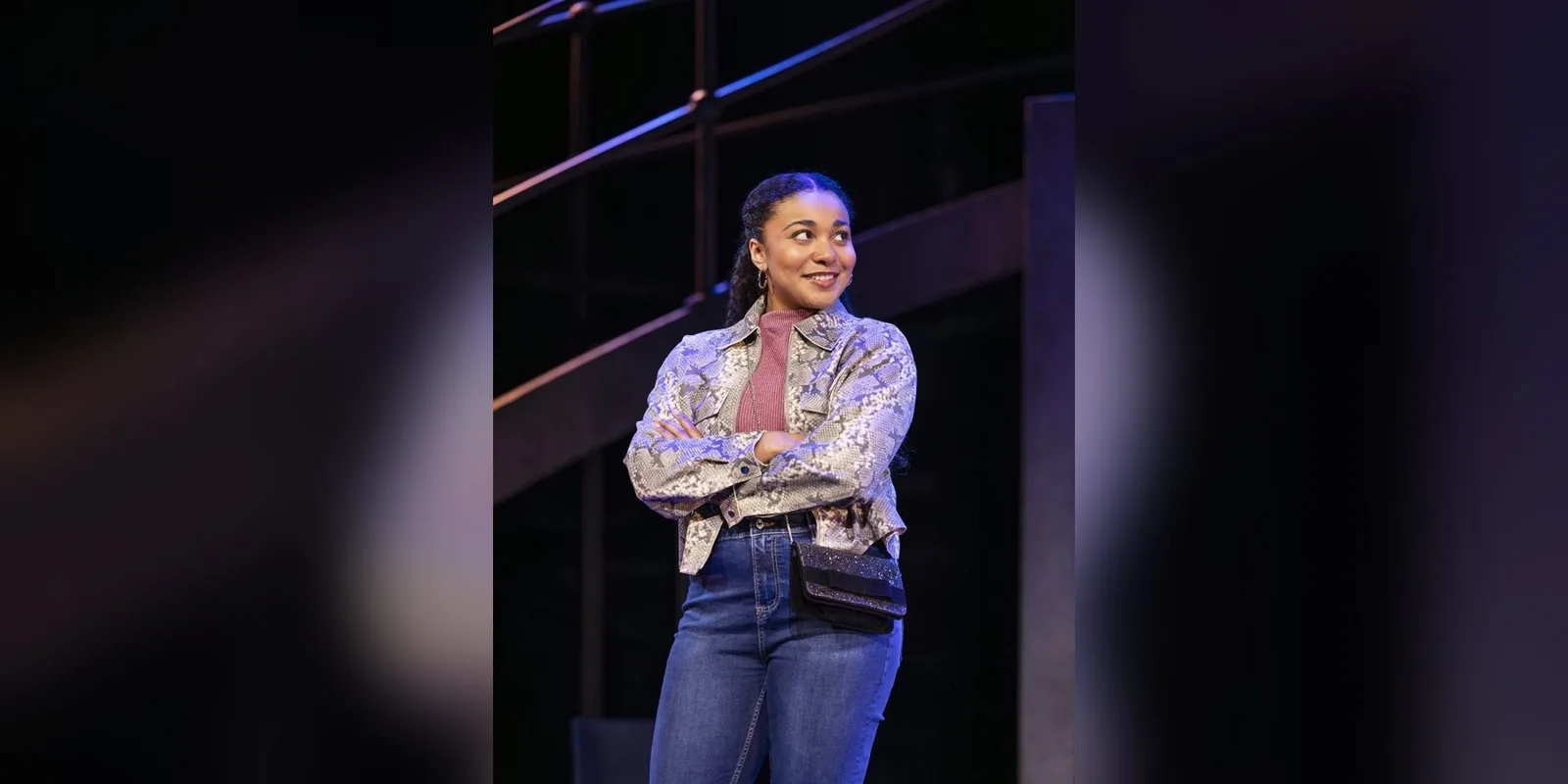 Grace Hodgett Young smiles with arms folded in a shiny patterned jacket and jeans in the 2026 production of James Graham's play
