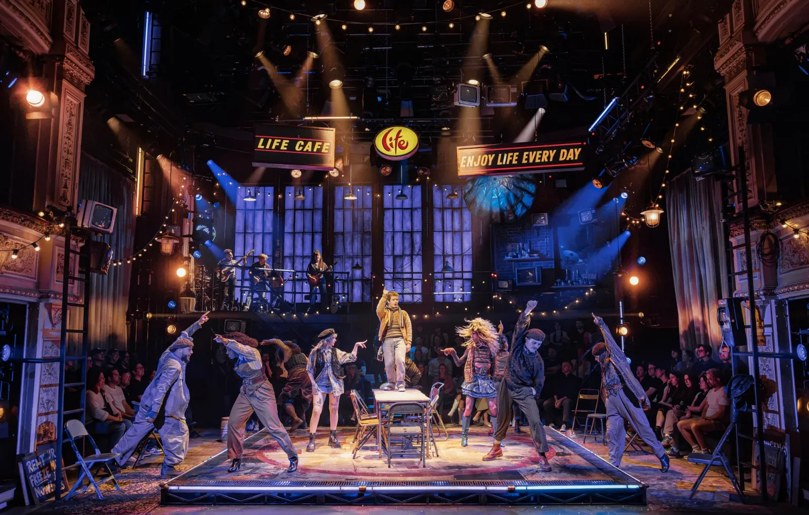 Gaten Matarazzo as Mark stands on a cafe table as the cast of Rent dance around him beneath a Life Cafe sign