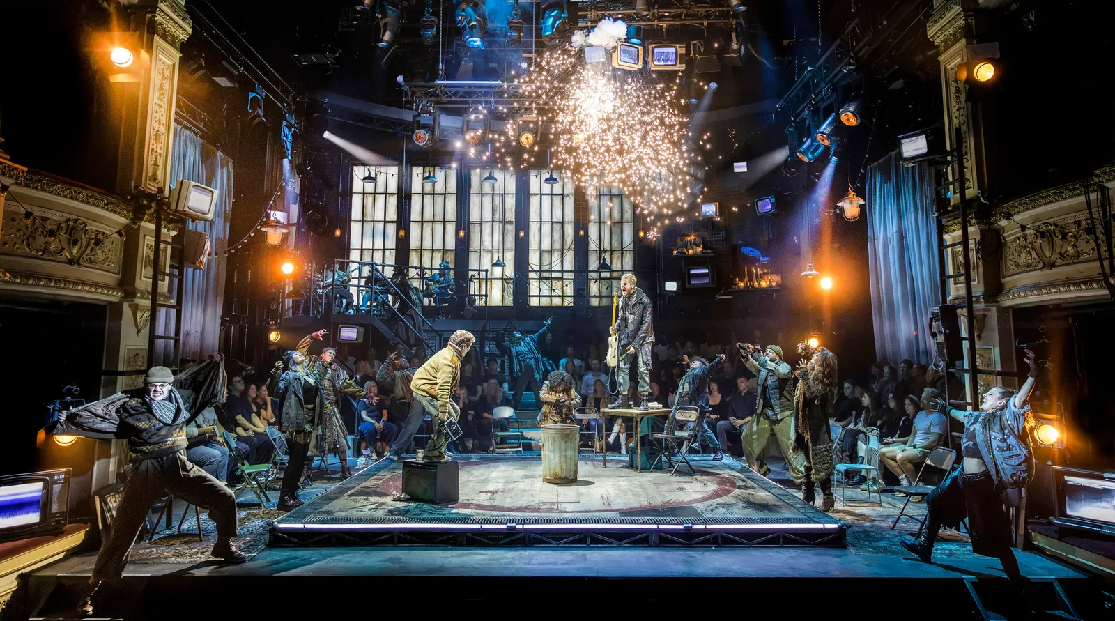 The full cast of Rent pose around a square stage as sparks shower down from the lighting rig