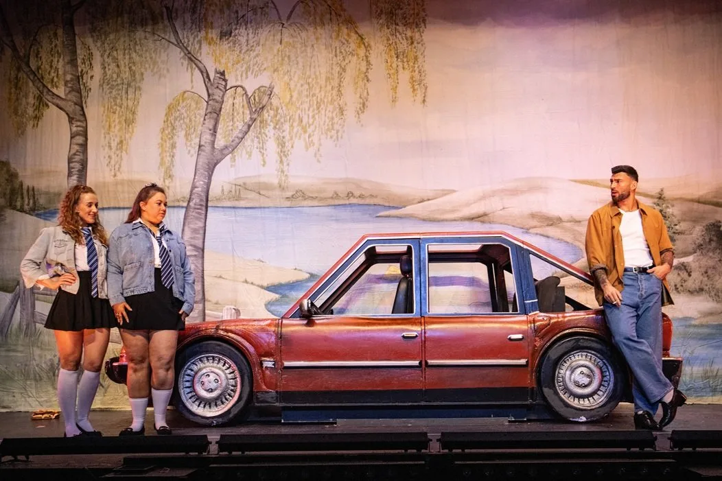 Two young women in denim jackets and a man in a tan jacket stand beside a red car on stage in the 2025 touring production of Andrea Dunbar's play