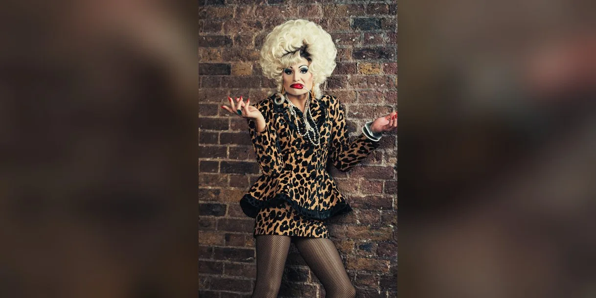 A drag performer in a huge curled platinum wig, leopard print suit and pearls stands against a brick wall with arms spread wide and a cigarette between red lips
