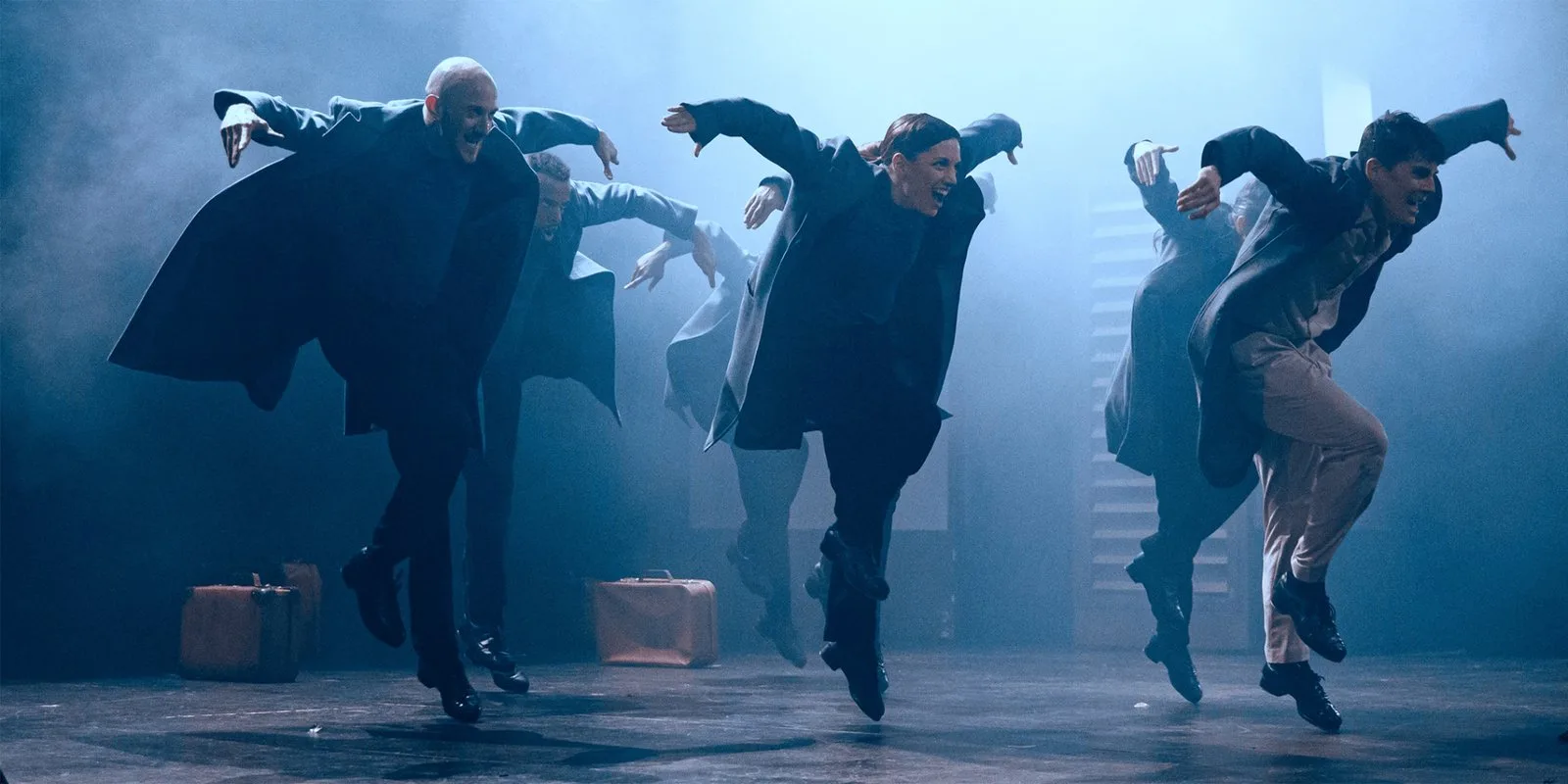Dancers in long dark coats leap in unison across a smoky, blue lit stage, arms flung out to the sides, with old suitcases at their feet