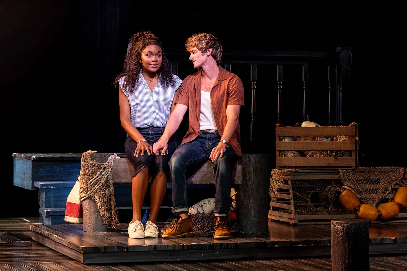 A young couple sit hand in hand on a wooden jetty bench beside lobster crates and fishing nets, the man turning towards the woman as she looks away