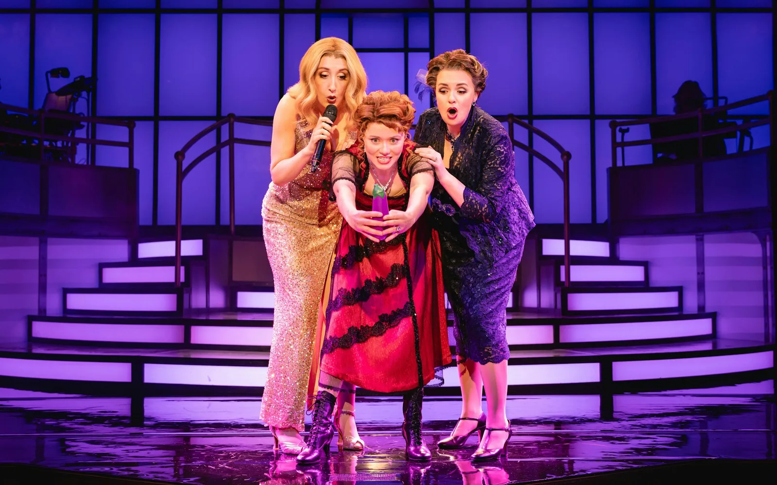 Three performers in Titanique sing together on a purple-lit staircase, one in a gold gown holding a microphone