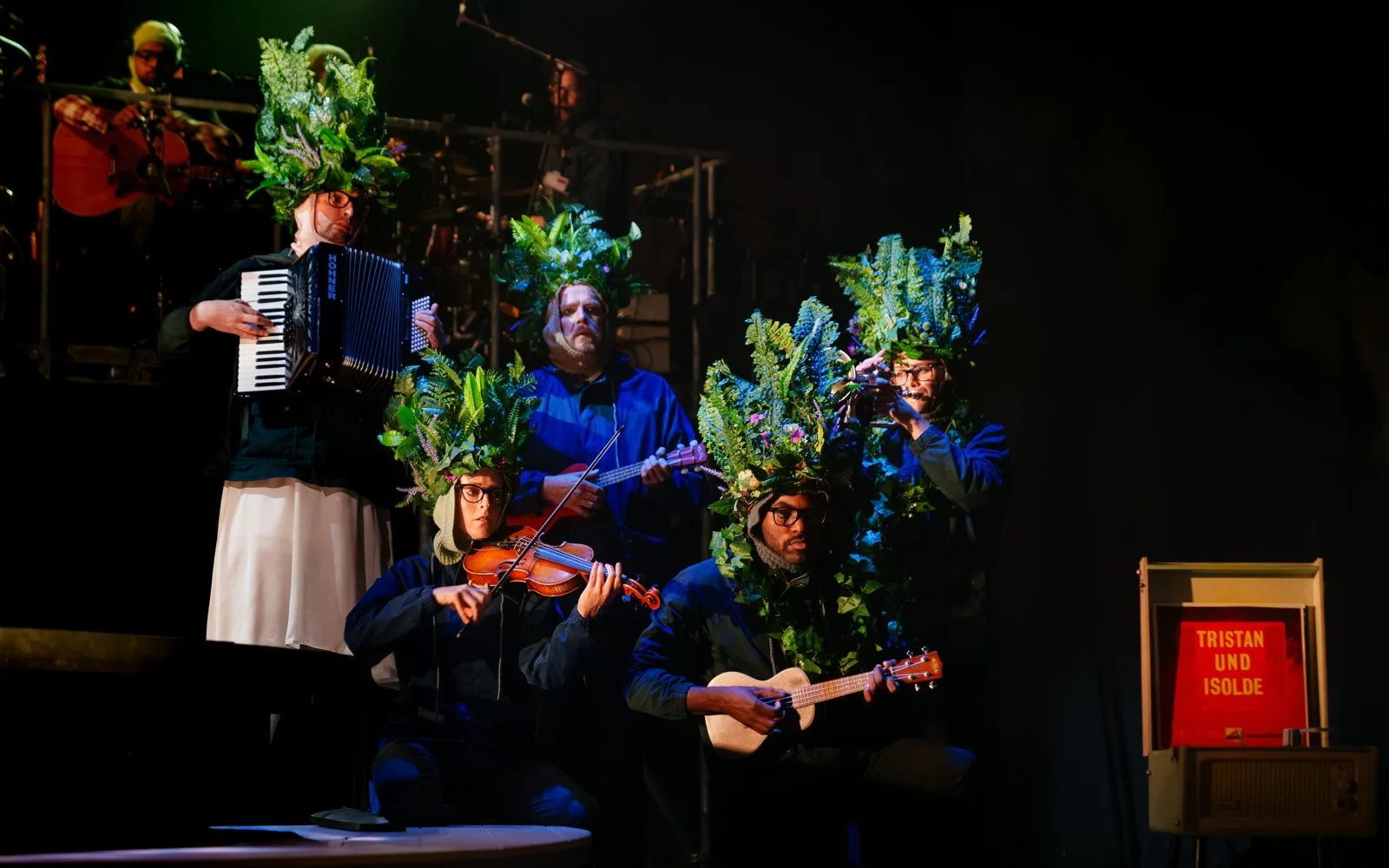 Musicians wearing headdresses of ferns and flowers play accordion, violin, ukulele and trumpet beside a vintage record player