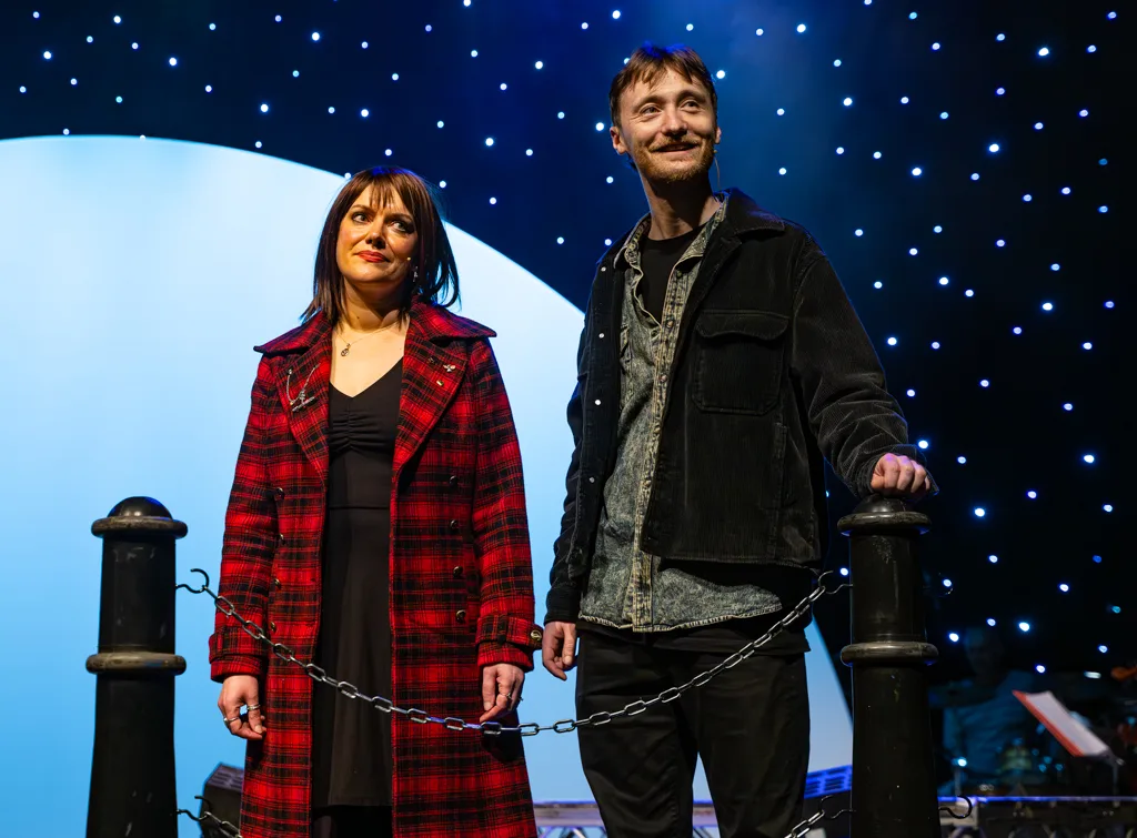 Two cast members of Under the Mersey Moon on a waterfront-style set with moon and starlit backdrop