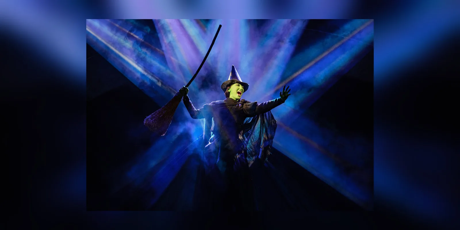 A green skinned figure in a dark hat and flowing cape raises one arm while holding a broom, lit by sweeping beams of blue light
