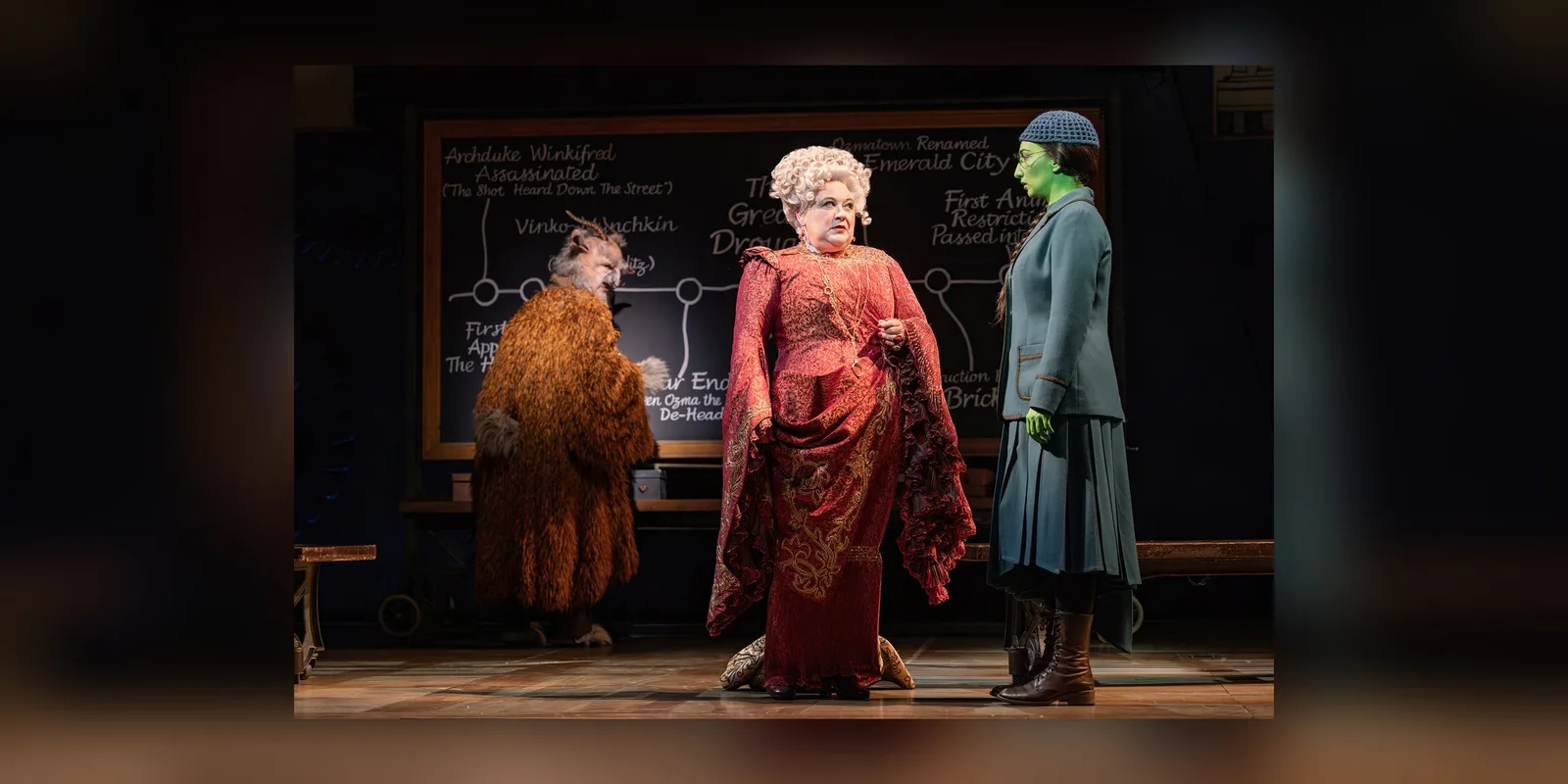 A woman in an ornate crimson gown and white curled wig stands before a chalkboard, between a goat in a shaggy coat and a green skinned student in a grey uniform