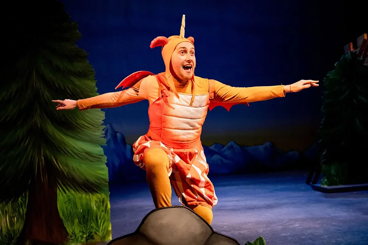 A performer in an orange dragon costume with wings and a single horn plays Zog, arms spread wide as if flying, in Zog and the Flying Doctors