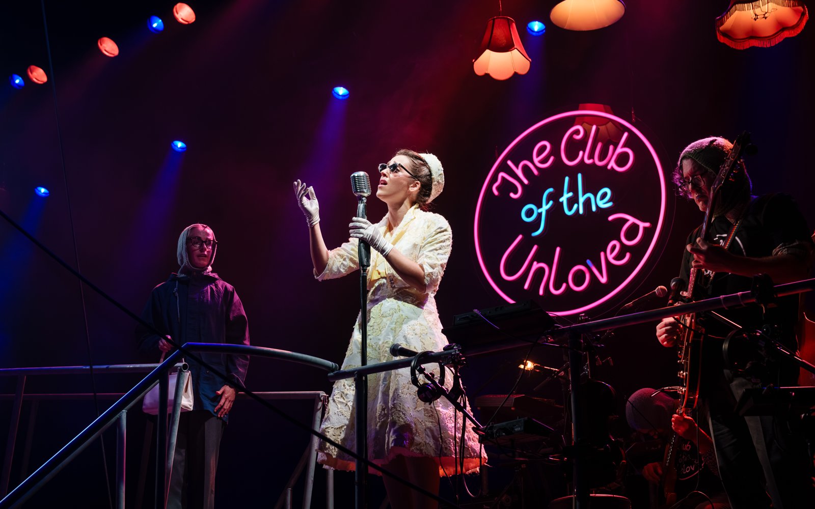 Stephanie Hockley as Whitehands singing at a vintage microphone beneath the neon Club of the Unloved sign, with Philippa Hogg and Pat Moran in Tristan & Yseult. Photo: Steve Tanner
